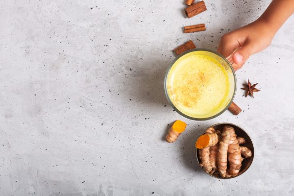 Traditional Indian Golden Turmeric Curcuma Milk On The Gray Concrete Background With Ingredients. Horizontal Orientation. Boys Hand Holds A Glass